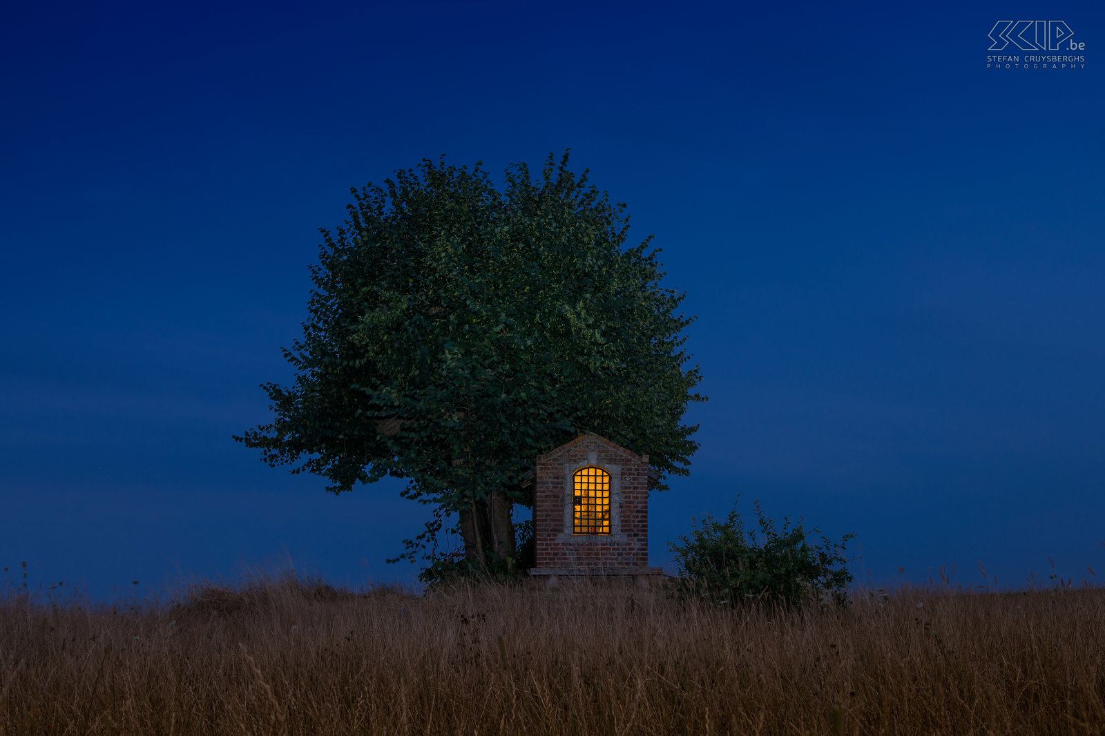 Hageland by night - Sint-Jozef kapel Onder een oude lindeboom midden in de velden van Sint-Pieters-Rode (Holsbeek) staat het Sint Jozefs kapelletje. Het eenvoudige kapelletje bestaat uit bakstenen en heeft een zandstenen omlijsting rondom de nis. Het dateert van het begin van de 19de eeuw. Deze foto werd gemaakt juli in het begin van het blauwe uur (het uur na zonsondergang). Stefan Cruysberghs